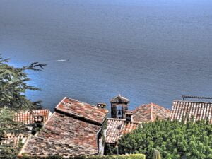 Vue plongeante sur Eze et la Méditerranée. On aperçoit une cloche sur le toit d'une maison bourgeoise et un bateau naviguant sur la mer au loin en contrebas